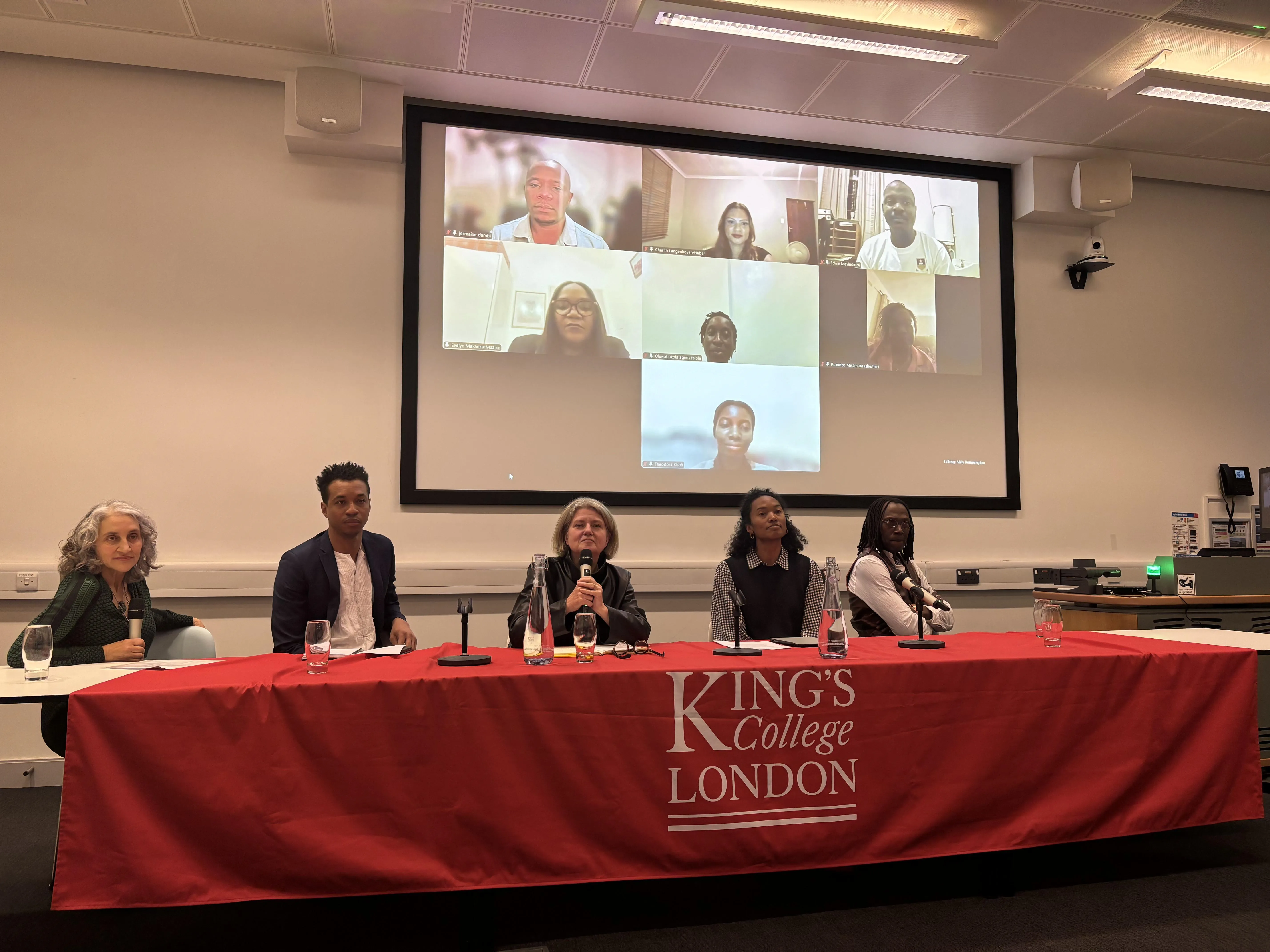 Panel table with five panelists seated. Large screen behind with 7 online panelists displayed. King's College London logo on red table cloth covering panel table