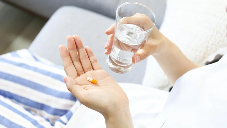A woman holding a pill in one hand and a glass of water in the other, preparing to take her medicine.