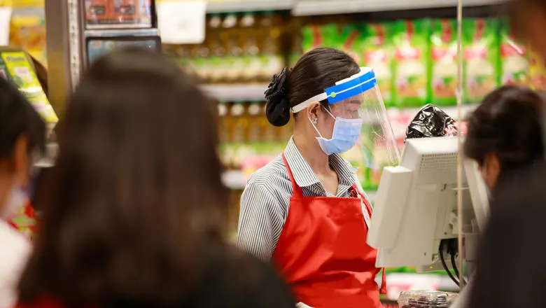 A female cashier wearing a covid facemask and visor works at a till. In the foreground, silhouettes of customers queuing.