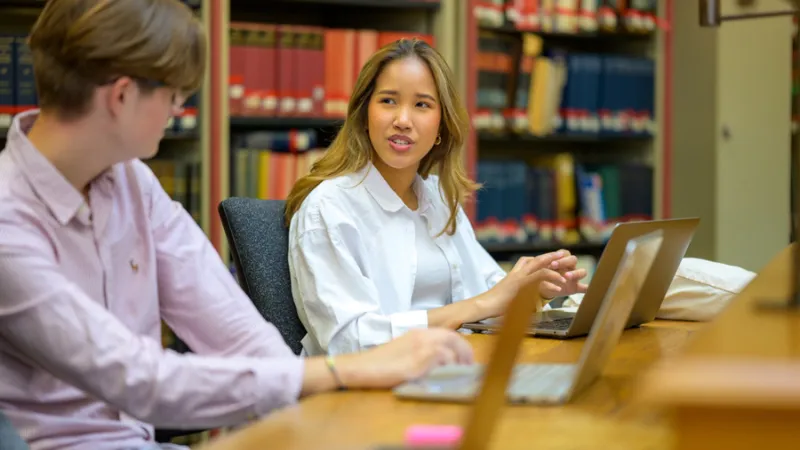 Two students in conversation with laptops in a library